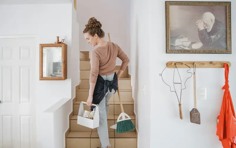 A cleaner carrying a caddy of supplies and a broom up the stairs of a home