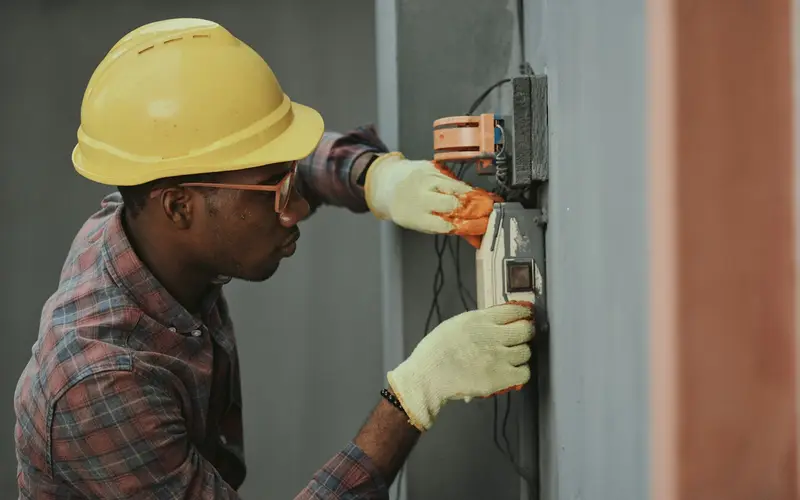 An electrician in a hard hat and gloves working on a wall panel