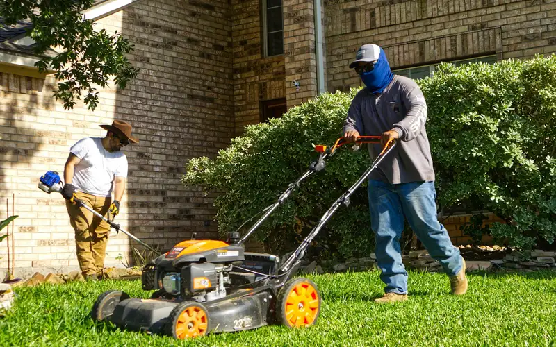 Two crew members mowing and trimming the lawn of a brick house