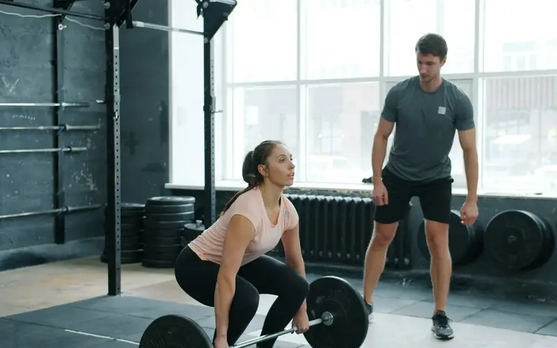 A trainer watching a client lift a barbell in a gym